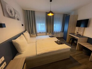 Una habitación de hotel con cama, escritorio, silla, televisor y ventana con cortinas.