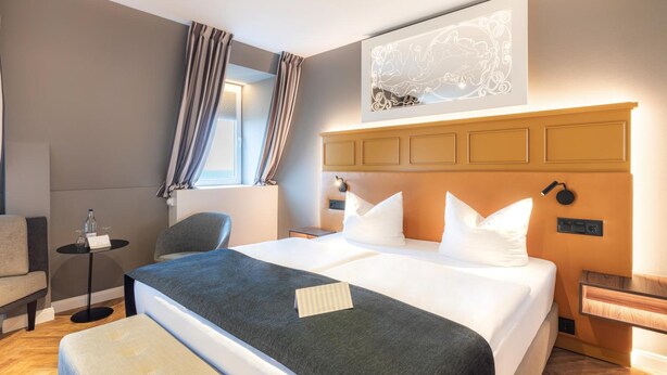 A hotel room with a large bed, a bedside table, a chair, a small table with a bottle and glasses, and a window with curtains. - Ringhotel Ostseehotel Ahlbeck (Heringsdorf)