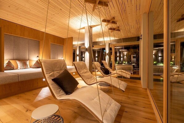 A modern lounge area with wooden flooring and ceiling, cushioned lounge chairs, and a seating area with a sofa and pillows. - Romantik Hotel Sackmann (Baiersbronn)