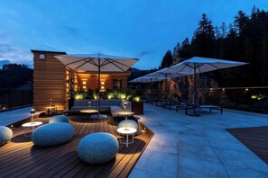 An outdoor seating area with round tables, benches, and umbrellas, surrounded by a wooden deck and a stone wall. - Romantik Hotel Sackmann (Baiersbronn)