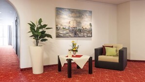 Un hall d’hôtel avec un tapis rouge à motifs, une grande plante en pot, une table sur laquelle est posé un vase de fleurs, un canapé et un tableau représentant un paysage urbain accroché au mur.