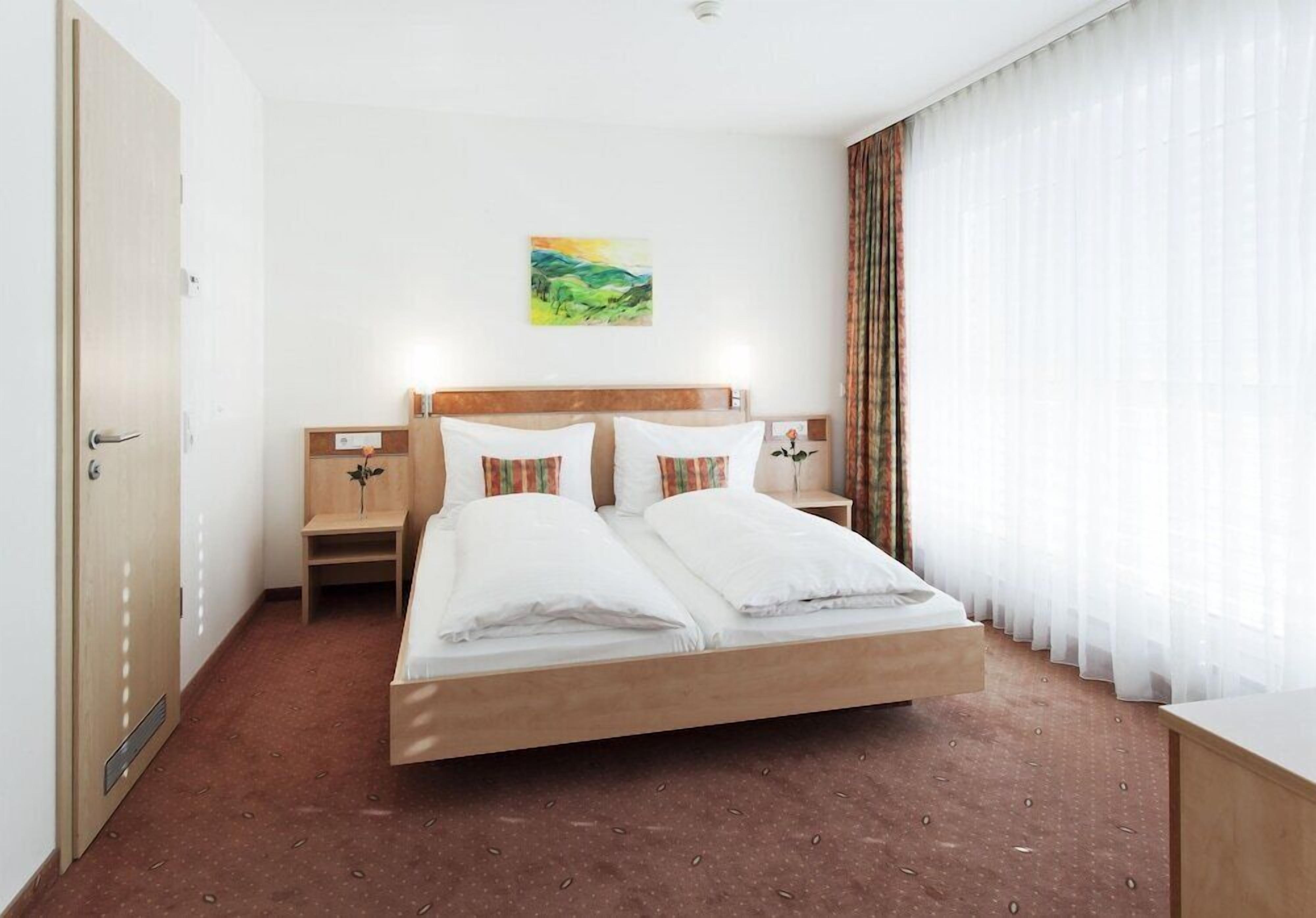 A hotel room with a bed, bedside tables, a nightstand, a painting on the wall, and a window with curtains.