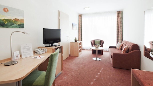 Une chambre d’hôtel équipée d’un bureau, d’une télévision, d’un fauteuil vert, d’un canapé marron et d’une petite table.