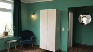 Una habitación con paredes verde azulado, un armario blanco, un sillón azul, una mesita redonda pequeña y una planta en maceta junto a la ventana.