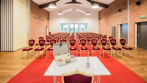 Un salón de conferencias con filas de sillas rojas, una mesa blanca con una maleta y una botella de agua, y paredes de ladrillo con grandes ventanales.
