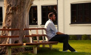Un uomo seduto su una panchina in un parco, con un edificio sullo sfondo.