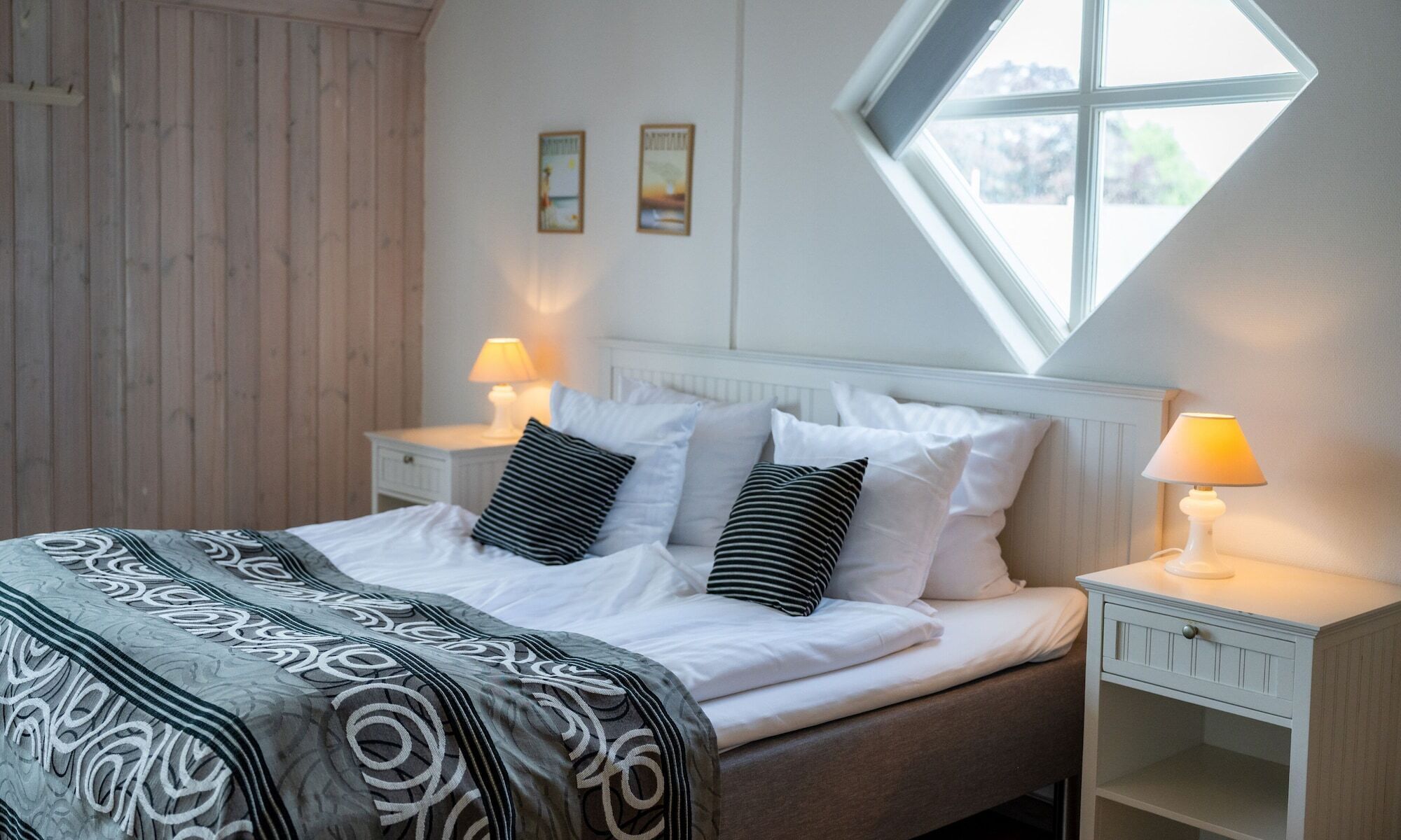 A bedroom with a bed, two bedside tables, a window, and two wall-mounted pictures.