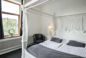 Una cama con dosel, cabecera blanca y ropa de cama gris, un sillón de cuero negro, una mesita de noche con lámpara y vista a un árbol por la ventana.