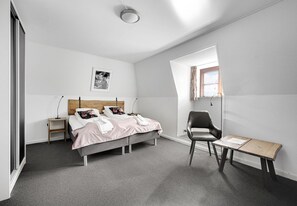 Un dormitorio con cama, mesitas de noche, una silla, una pequeña mesa y una ventana.
