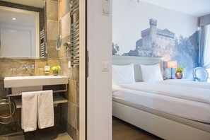 A hotel room with a bathroom featuring a sink, mirror, and towel rack. The bedroom has a bed, nightstand, lamp, and a wall-mounted mirror. - Boutique Business Hotel La Tureta (Bellinzona)