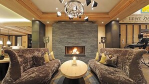 A cozy living room with a fireplace, two patterned sofas, a round coffee table, and a chandelier. - Wellness Spa Hotel Ermitage (Saanen)