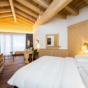 A hotel room with a wooden ceiling, a bed, a desk with a chair, a small table with a vase, and a window with curtains. - Wellness SPA Hotel Ermitage (Saanen)