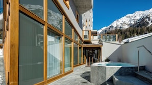 A modern building with large glass windows, a stone patio, and a hot tub. - TOP Hotel Sarain Active Mountain Resort (Lantsch-Lenz)