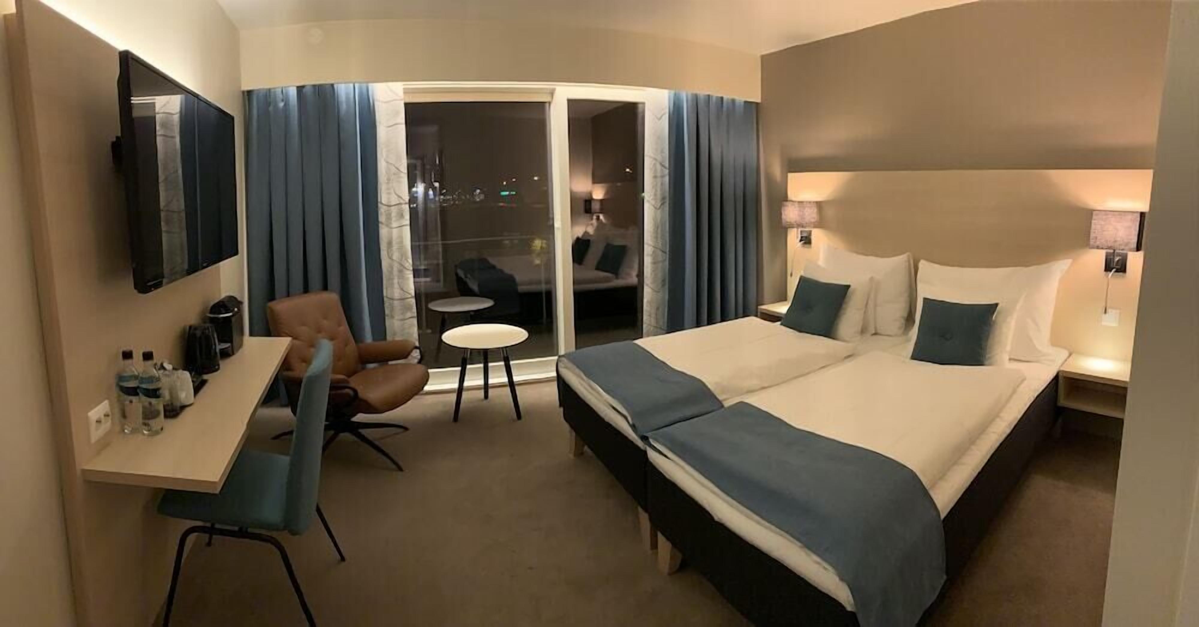 Ein Hotelzimmer mit einem Bett, einem Schreibtisch, einem Sessel, einem kleinen Tisch und einer Schiebetür, die auf einen Balkon führt.