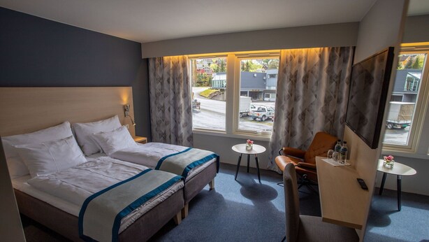 Une chambre d’hôtel avec un grand lit, un bureau, une chaise et une petite table. Les fenêtres sont dotées de rideaux et offrent une vue sur la rue.