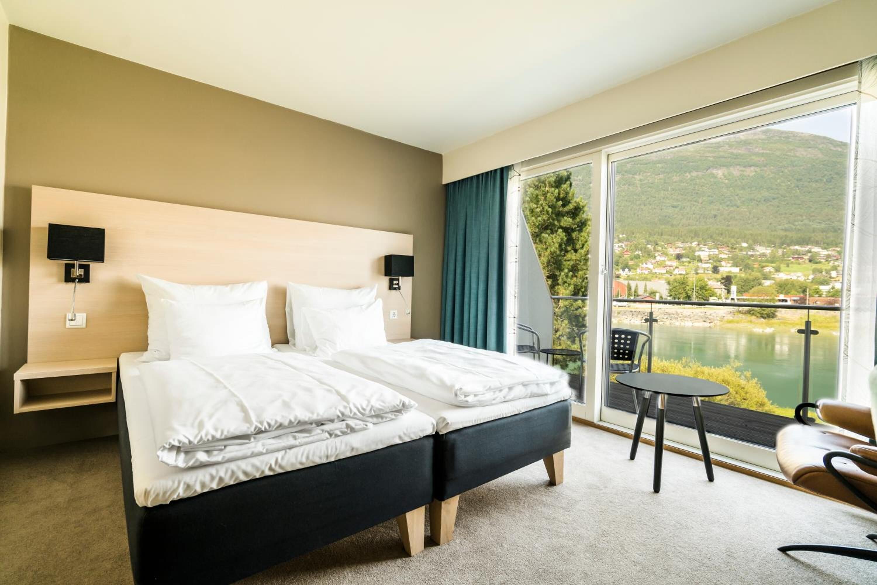 A hotel room with a large bed, a small table, and a chair. There is a balcony with a view of a lake and mountains.