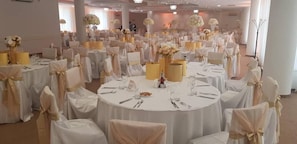 A banquet hall with round tables, white tablecloths, and chairs with gold sashes. - Arpesos (Kardzali)