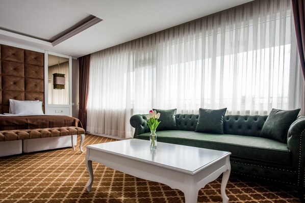 Habitación de hotel con cama, sofá verde, una mesita de café blanca y un ventanal con cortinas traslúcidas.