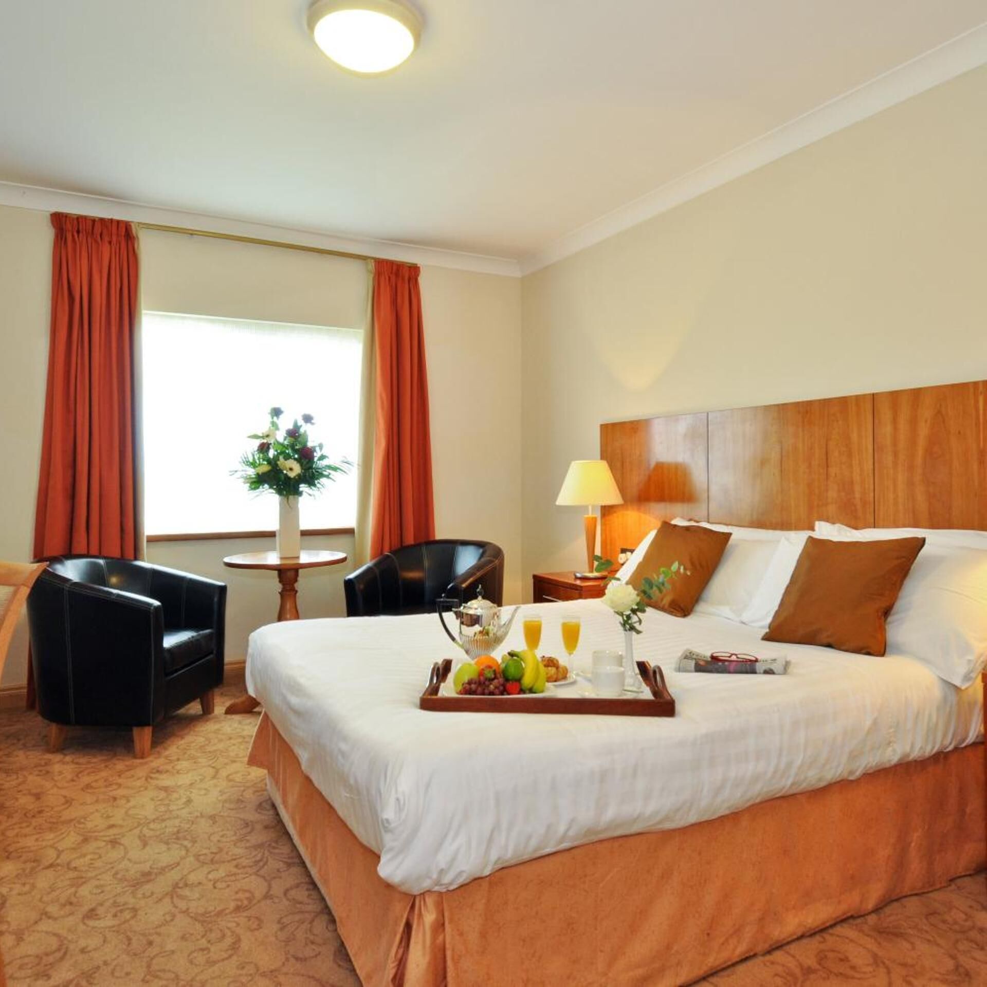A hotel room with a large bed, a small table with a vase, two chairs, and a window with curtains.