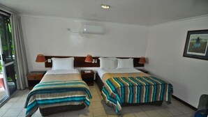 Habitación de hotel con dos camas, cada una con cubrecama a rayas. Tiene una ventana con cortinas, un aire acondicionado de pared y un cuadro enmarcado.