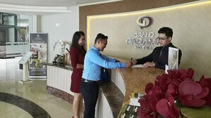 Uma receção com o letreiro do Grand Paramus Hotel, uma mulher de vestido vermelho e um homem de camisa azul apertando as mãos.