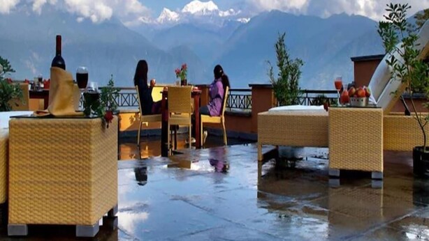 A rooftop terrace with wicker furniture, a table with a bottle and glasses, and a view of snow-capped mountains. - Club Mahindra Chumbi (Gyalshing)