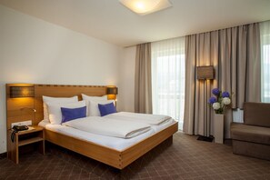 Una habitación de hotel con cama de madera, una mesita de noche, un escritorio, una silla y un amplio ventanal con cortinas.