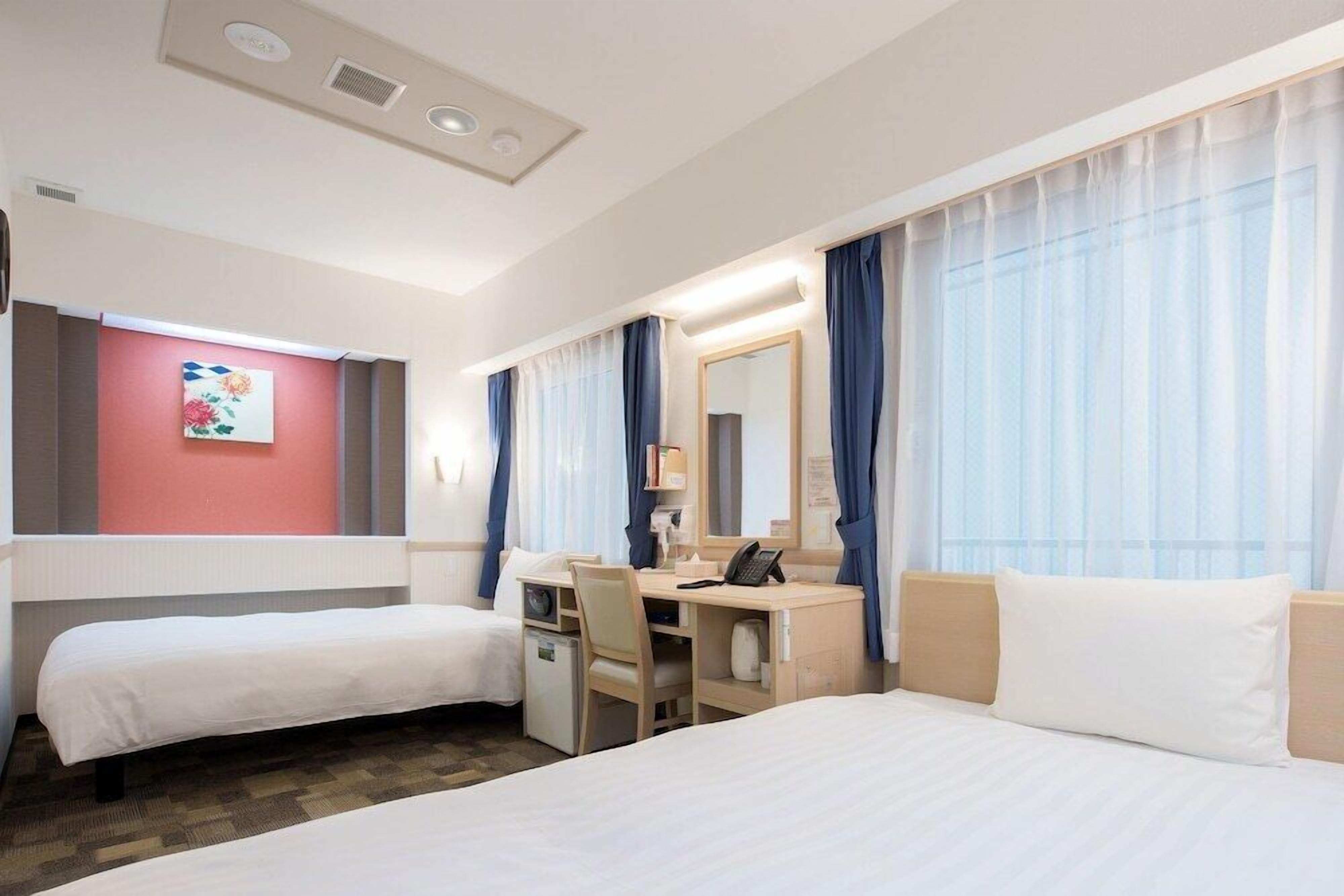 A hotel room with two single beds, a desk with a chair, a small desk lamp, and a telephone.