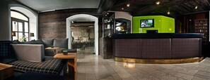 A lobby with a checkered sofa, a reception desk, and a television screen. - Hotel Jakob (Fuschl am See)