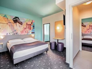 Une chambre d’hôtel moderne avec un lit, un grand tableau abstrait, une petite table avec deux tabourets et un miroir.