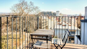 A balcony with a wooden table and bench, overlooking a cityscape with buildings and trees.