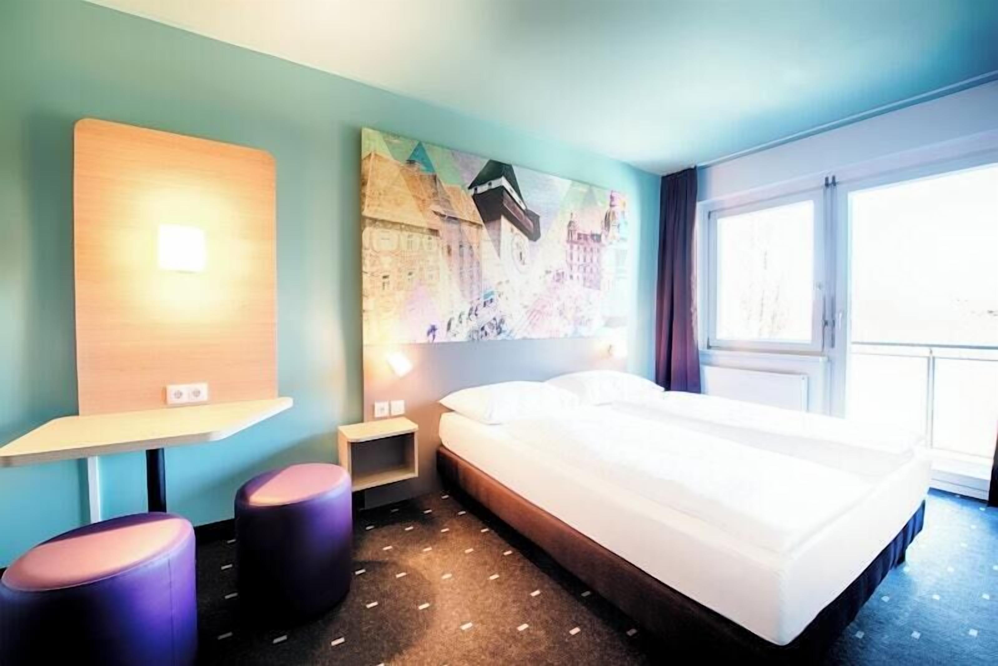 Ein Hotelzimmer mit einem Bett, einem Schreibtisch, zwei Stühlen, einem Nachttisch und einem großen Fenster.