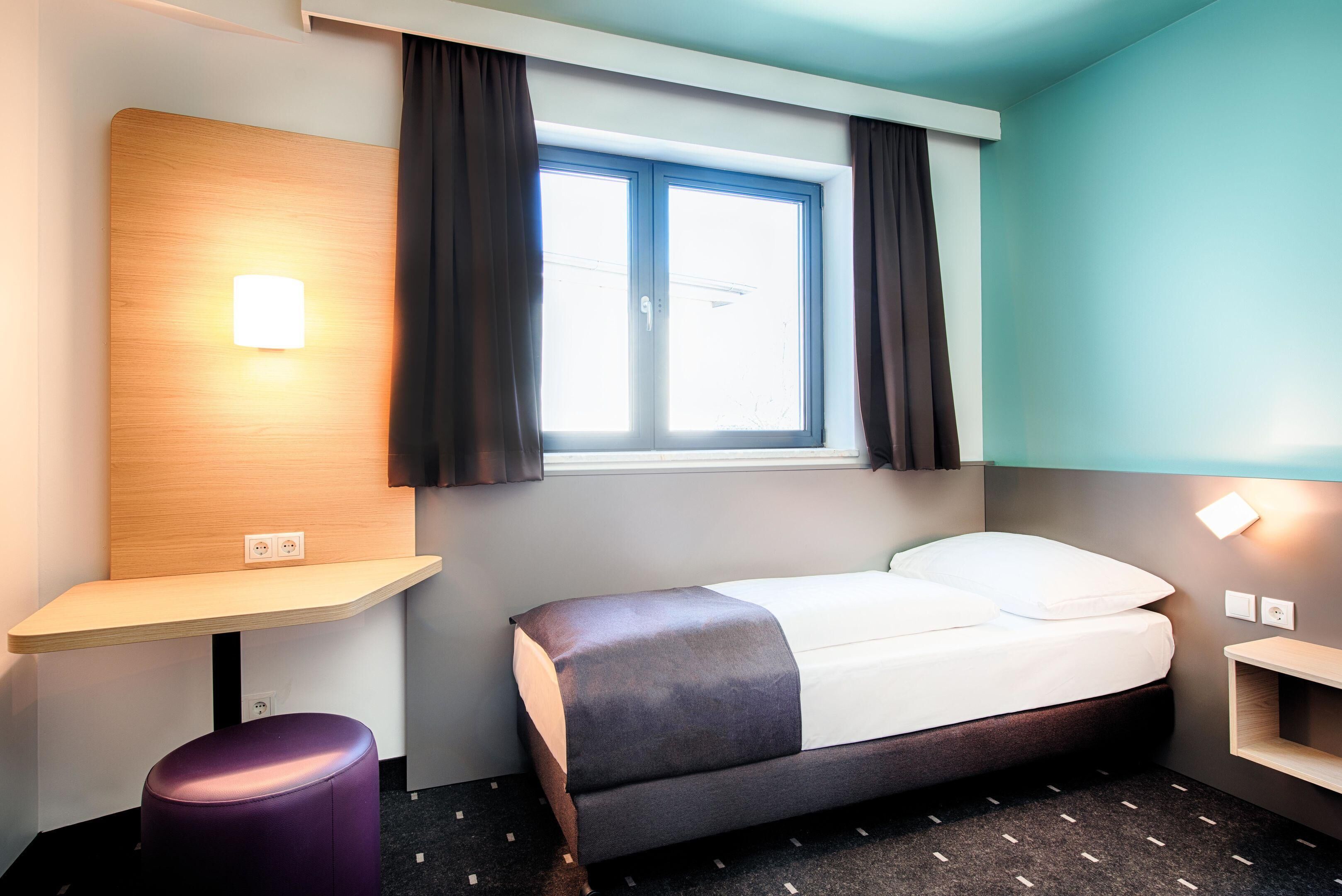 A hotel room with a bed, a desk, a chair, a window with curtains, and a wall-mounted light.