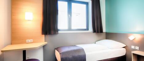 A hotel room with a bed, a desk, a chair, a window with curtains, and a wall-mounted light.