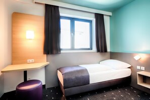 Habitación de hotel con cama, escritorio, silla, ventana con cortinas y una lámpara de pared.