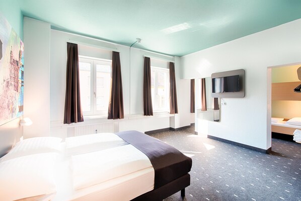 Una habitación de hotel con una cama, un televisor montado en la pared y un ventanal con cortinas.