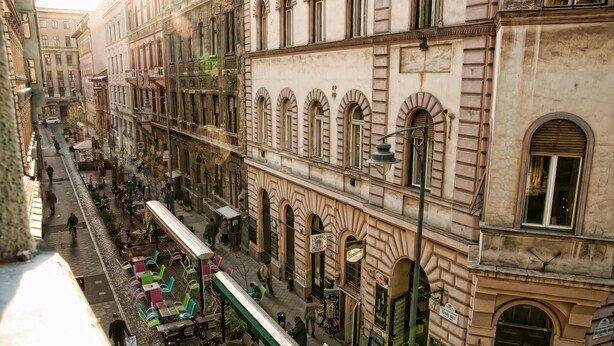 Une vue d’une rue où l’on aperçoit un bâtiment historique, avec un café au rez-de-chaussée, des tables à l’extérieur et une ligne de tramway.