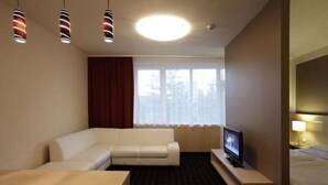 A hotel room with a white sofa, a small TV on a wooden stand, a bed with white bedding, a window with curtains, and a ceiling light. - Laterum Conference And Wellness (Pecs)
