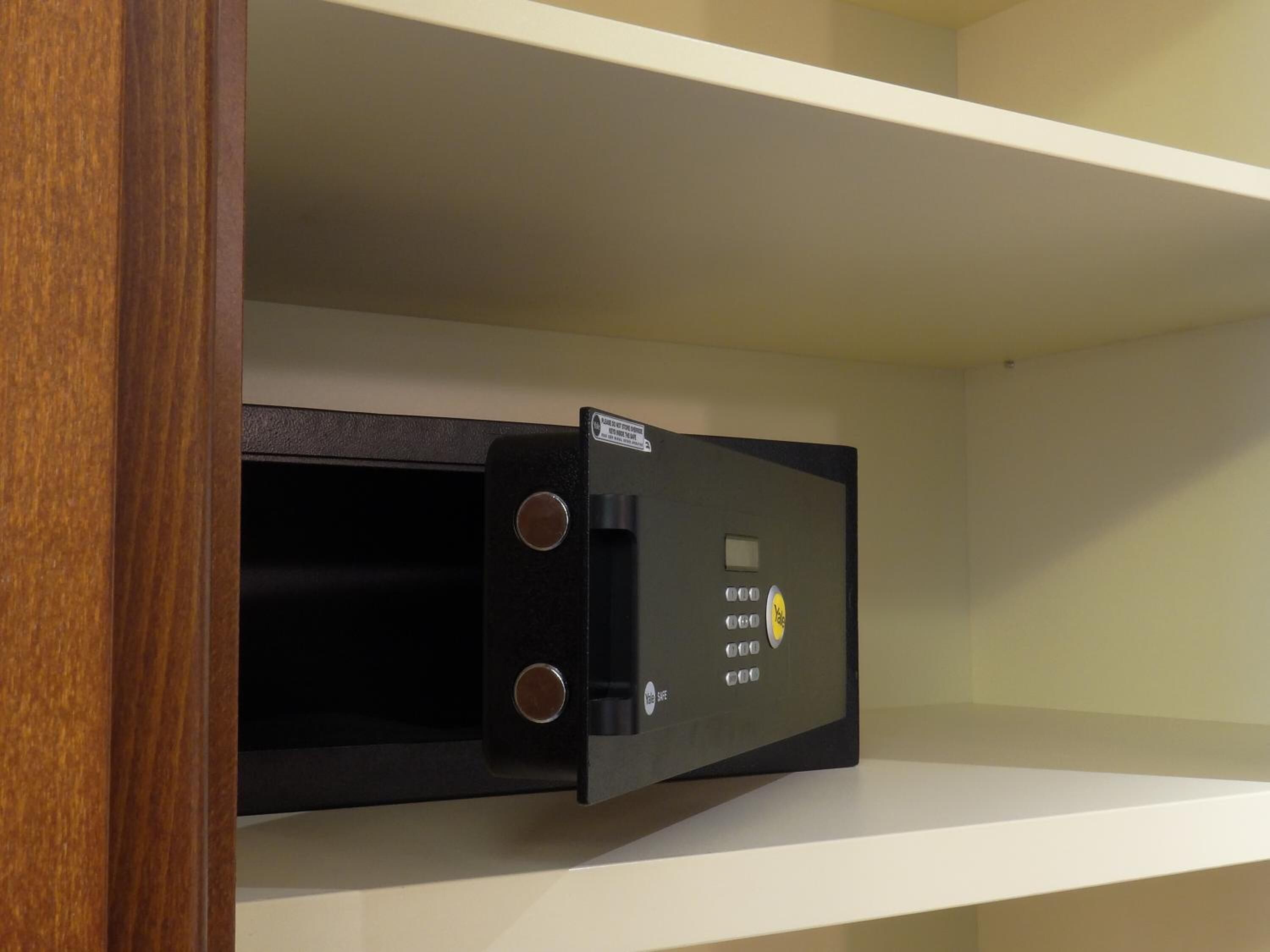 A black safe with a Yale lock, installed in a wooden cabinet with a white shelf above it.