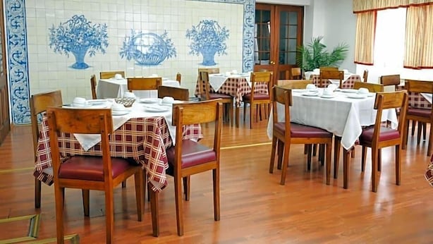 A dining area with wooden chairs and tables, a tiled wall with blue floral patterns, and a window with curtains. - Residencial Sao Francisco (Horta)