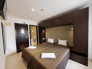 A hotel room with a large bed, a wardrobe, a bathroom, and a door with an exit sign. - Residencial Sao Francisco (Horta)