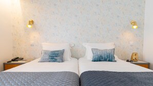 Una cama doble con una colcha gris acolchada y dos almohadas, una blanca y otra con un motivo azul. Hay dos mesitas de noche, una con una lámpara y un teléfono, y la otra con un objeto decorativo.