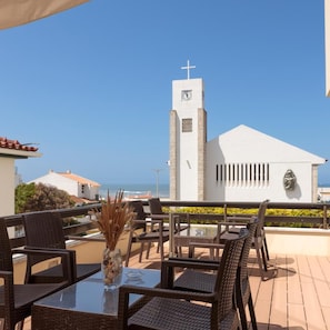An outdoor seating area with wicker chairs and a glass table, overlooking a white building with a cross on top. - Residencial Miramar (Marinha Grande)