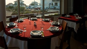 A dining area with tables set for two, large windows with a view of buildings and parked cars, and a red tablecloth. - Residencial Miramar (Marinha Grande)