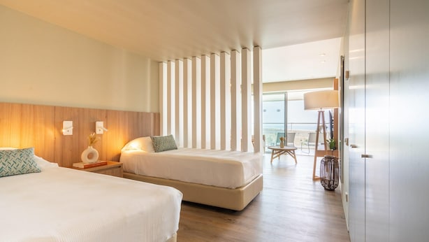 Une chambre d’hôtel avec deux lits, une tête de lit en bois, une grande fenêtre avec des rideaux, une table de chevet avec une lampe et une vue sur la plage.