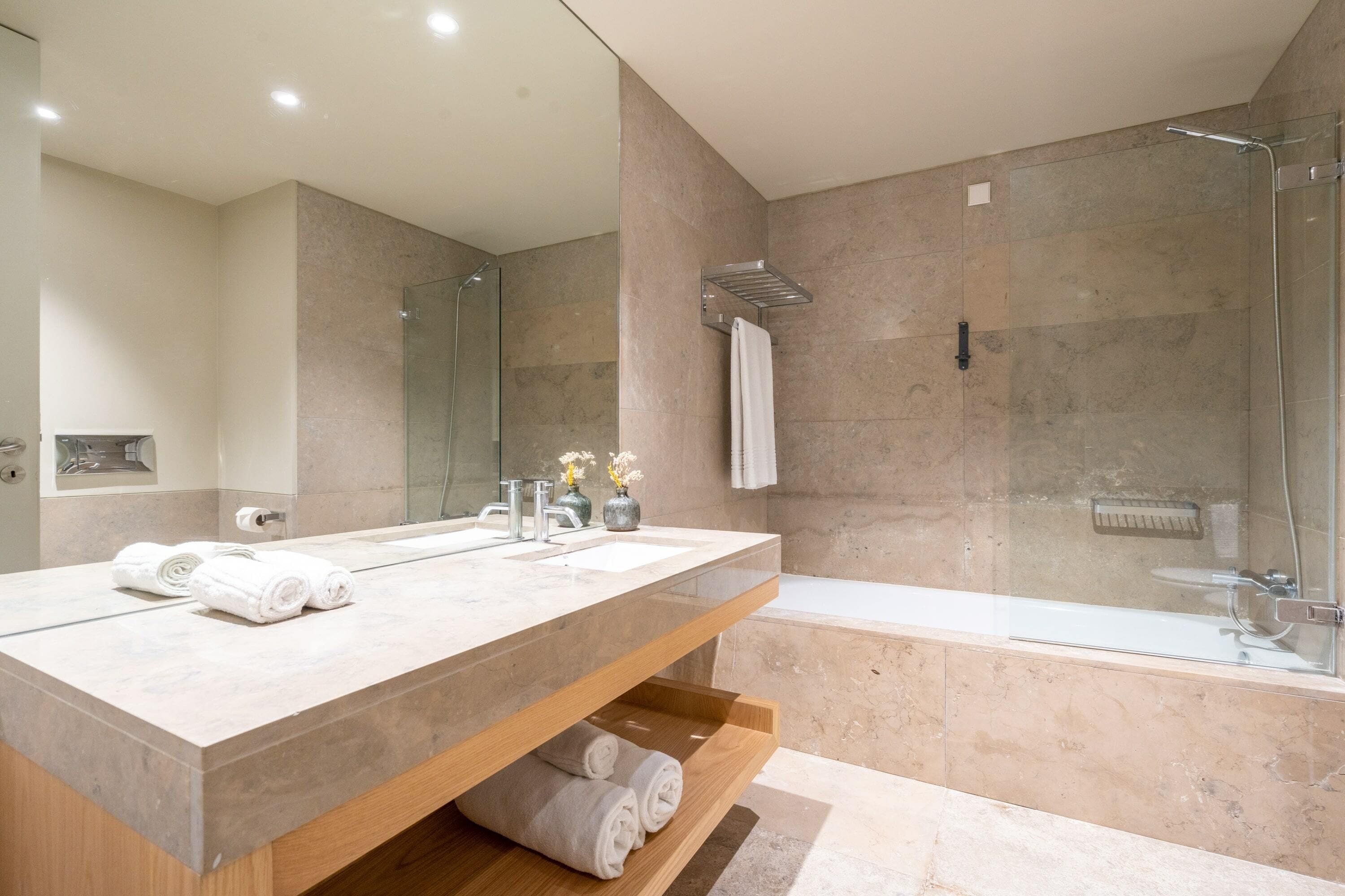 Un baño moderno con un espejo grande, dos lavamanos, una ducha y un estante de madera con toallas.
