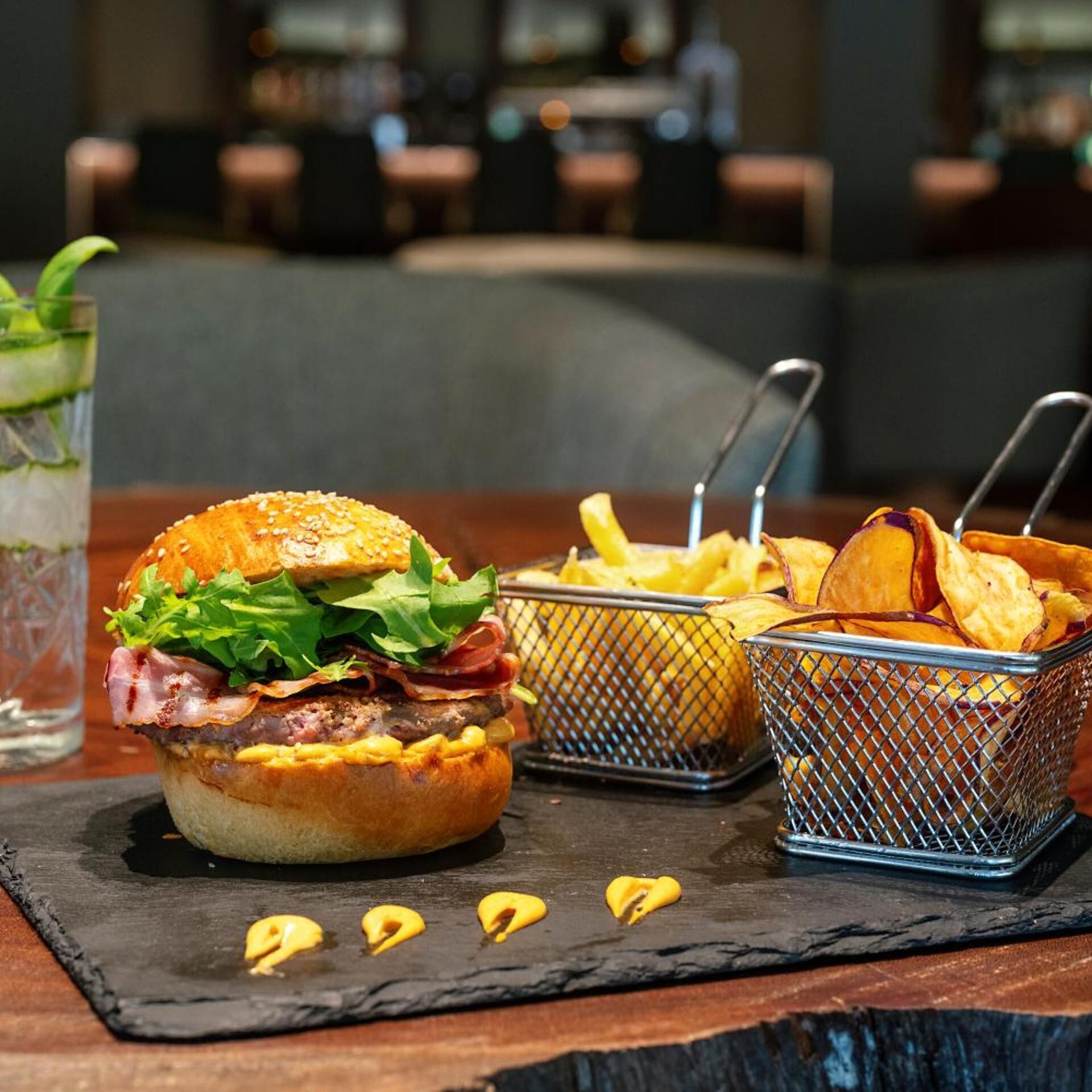 A burger with lettuce and bacon, two baskets of fries, and a glass of a beverage with a mint garnish on a slate board.