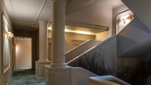 A hallway with a blue carpet, white columns, a staircase with a railing, and wall-mounted lighting. - Grande Hotel Lisboa (Sao Pedro do Sul)