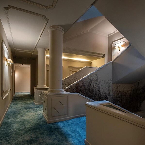 Un pasillo con alfombra azul, columnas blancas, una escalera con barandilla e iluminación en la pared.