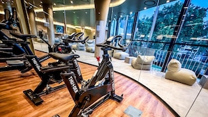 A modern gym with stationary bikes, a wooden floor, and large windows offering a view of the outdoors. - Grande Hotel Lisboa (Sao Pedro do Sul)